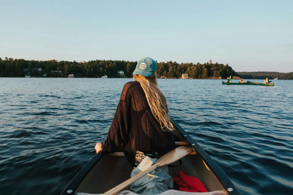 A person in a cap paddling a canoe on a lake at sunset